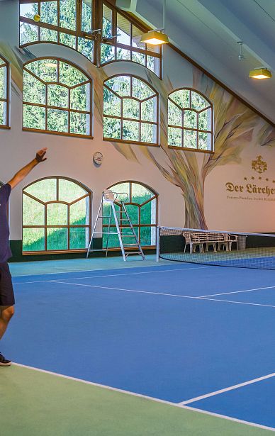 Ein Tennisspieler in sportlicher Kleidung schlägt den Ball auf einem Hallenplatz mit mehreren großen Fenstern und blauen Tennisplätzen auf.