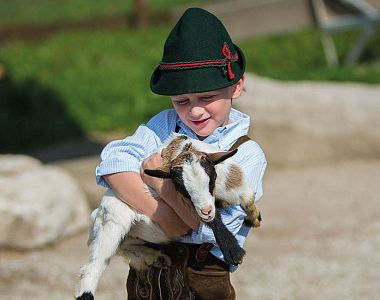 A young boy in Bavarian lederhosen and hat holds a small goat outdoors on a sunny day, set against a backdrop of green grass and rock formations.